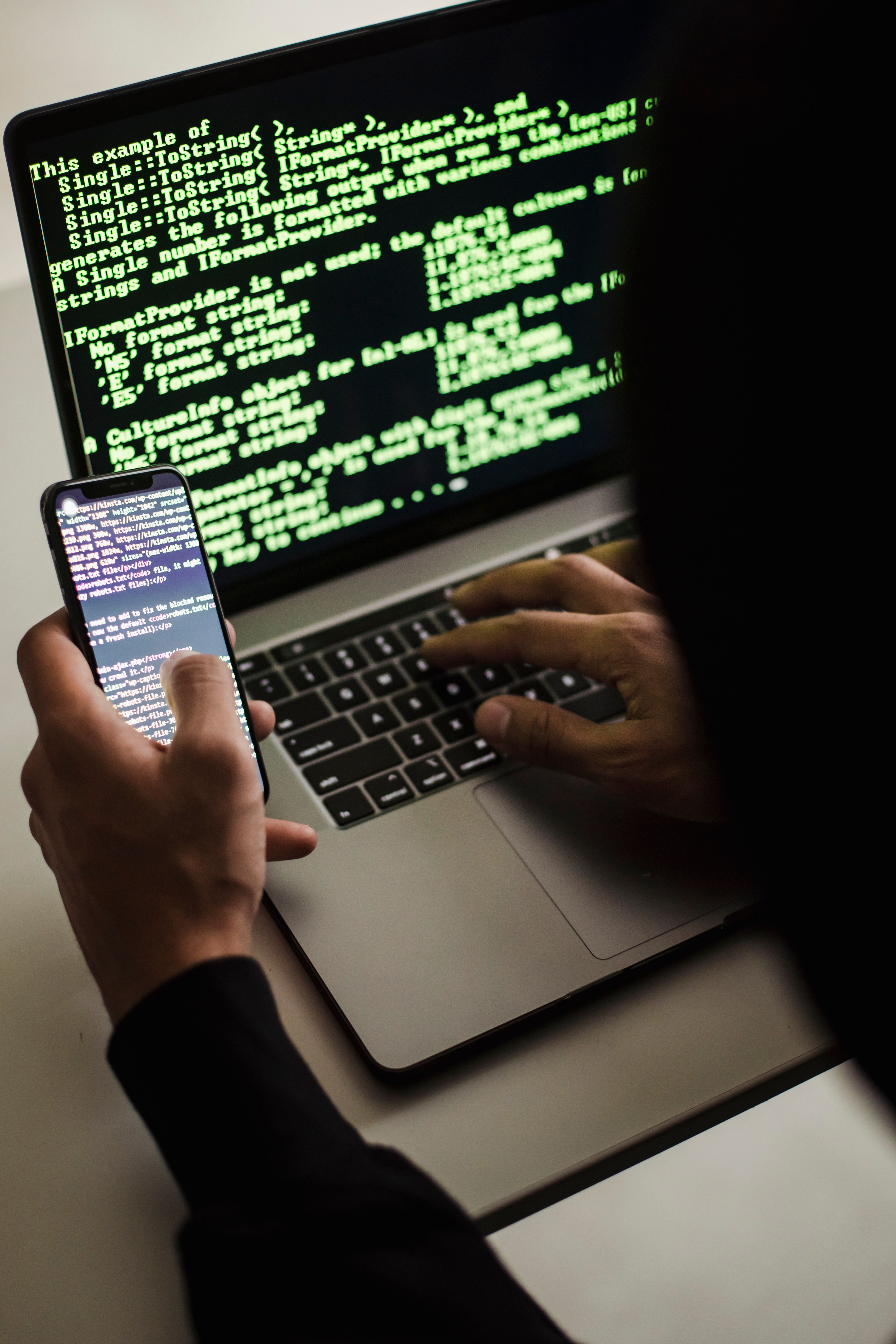 Anonyme Person in dunklem Kapuzenpullover arbeitet mit Laptop und Smartphone – Symbol für Cyberangriffe und Phishing-Bedrohungen (Sora Shimazaki via Pexels)